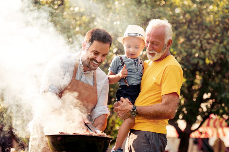 A grandfather in a yellow shirt holds a toddler wearing a straw hat and suspenders while a younger man in an apron grills meat on a charcoal barbecue outdoors, with smoke rising and green trees in the background