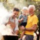 A grandfather in a yellow shirt holds a toddler wearing a straw hat and suspenders while a younger man in an apron grills meat on a charcoal barbecue outdoors, with smoke rising and green trees in the background