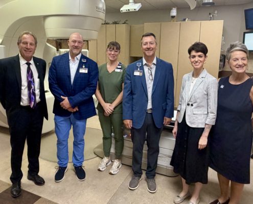Un grupo de seis profesionales médicos posan para una foto en un hospital, de pie frente a un gran escáner de resonancia magnética, con sus credenciales y uniformes quirúrgicos.