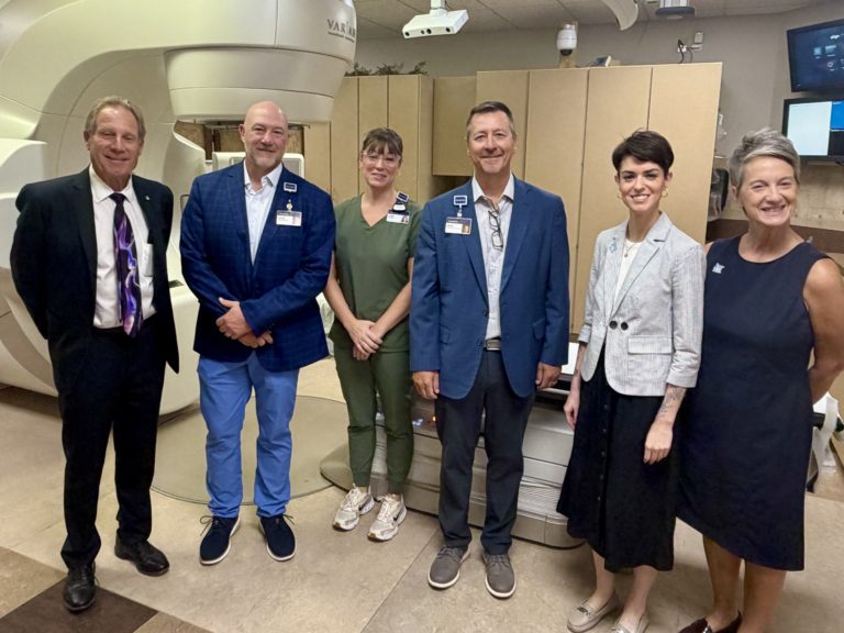 Group of six medical professionals posing for a photo in a hospital, standing in front of a large MRI scanner with badges and scrubs.