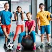Four children in colorful gym attire pose in a bright indoor gym with a soccer ball, dumbbells, and a stability ball nearby.