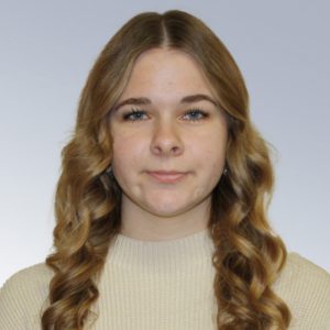Portrait of a young woman with long wavy brown hair wearing a beige knit sweater, against a light gray background.