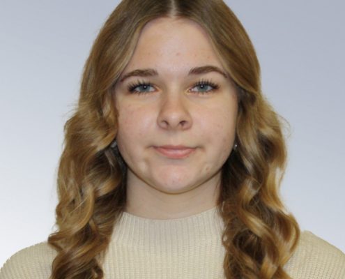 Portrait of a young woman with long wavy brown hair wearing a beige knit sweater, against a light gray background.
