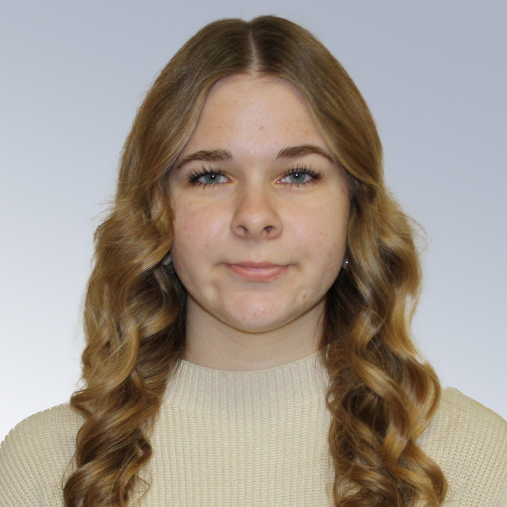 Portrait of a young woman with long wavy brown hair wearing a beige knit sweater, against a light gray background.